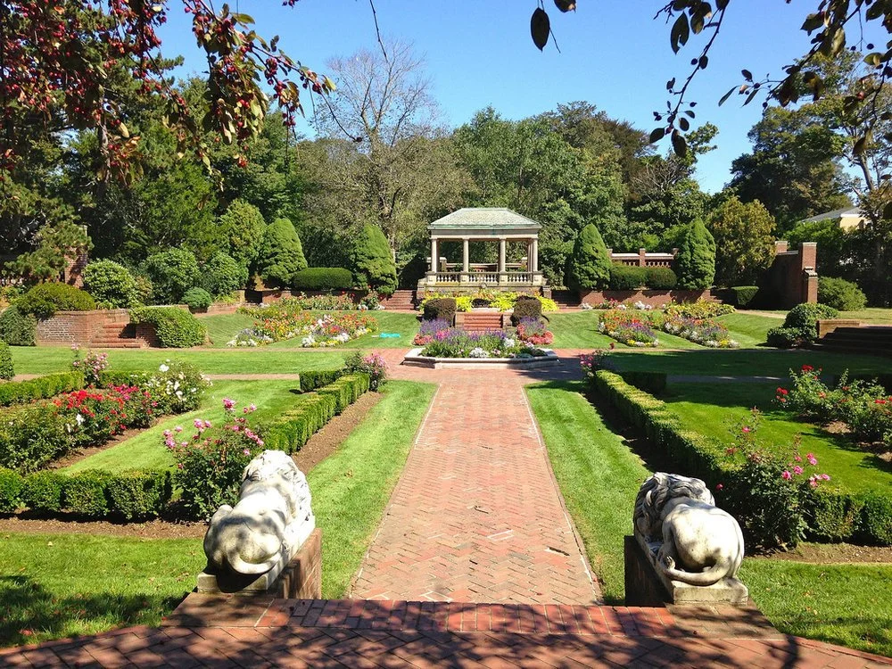 The Italian Garden at Lynch Park in Beverly's Cove neighborhood, with a columned pergola, marble lion statues, and formal rose beds under a blue sky.