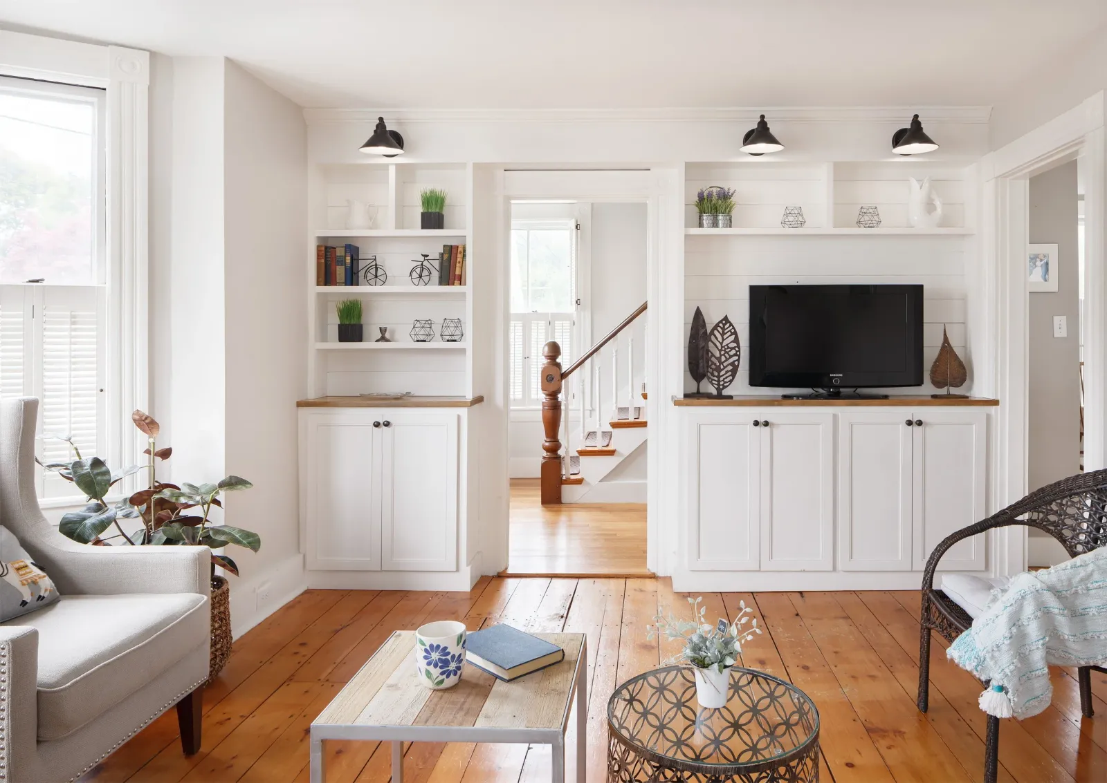 A bright staged living room with white built-ins, wide pine floors, and a staircase beyond